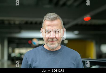 Confident mature man with gray hair and beard looking directly at the camera indoors against a softly blurred urban background. Stock Photo