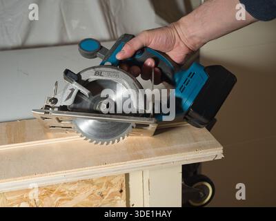 Hand Using Cordless Circular Saw to Cut Plywood; Close-up of a worker's hand holding and operating a battery-powered circular saw to cut a wooden plan Stock Photo