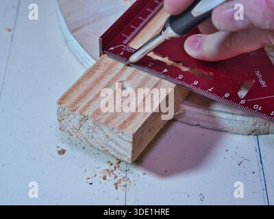 Carpenter Marking Wood Plank with Rafter Square; Close-up of a carpenter using a red precision rafter square and pencil to measure and mark a line on Stock Photo