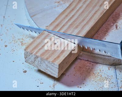 Handsaw blade cutting raw pine wood on workbench; Macro shot of a serrated hand saw blade starting a cut into a thick pine wood plank on a messy workb Stock Photo
