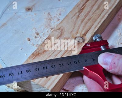 Woodworker Measuring Plank with Marking Gauge; Close-up of a carpenter using a red precision marking gauge and ruler to measure and mark raw wooden pl Stock Photo