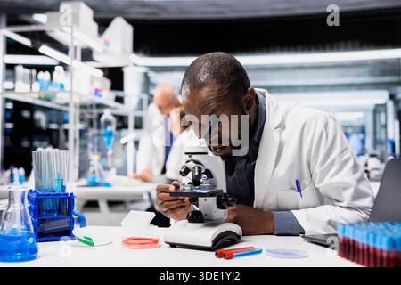 Materials scientist in lab checks product surface for tiny defects using microscope. African american man in research facility ensures manufacturing standards, examines small parts with optical device Stock Photo