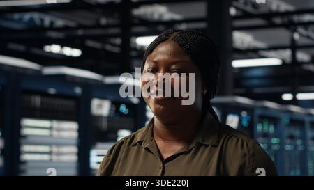 Portrait of cheerful data center engineer conducting security audits to protect against cyber threats. Jolly worker walking in server room, ensuring compliance with industry standards, camera B Stock Photo