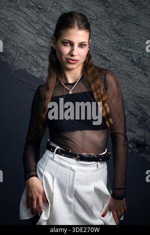 This image showcases a stylish young woman with long, wavy brown hair, confidently posing in a sheer black long-sleeved top paired with high-waisted w Stock Photo