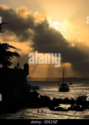Dramatic golden-hour sunset with sunbeams over the ocean, boats and coastal fort silhouette. Taken on Morro de São Paulo, Bahia, Brazil. Stock Photo