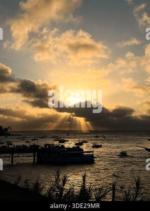 Golden-hour sunset with sunbeams streaming through dramatic clouds over the sea, with boats anchored across the bay near a pier Stock Photo
