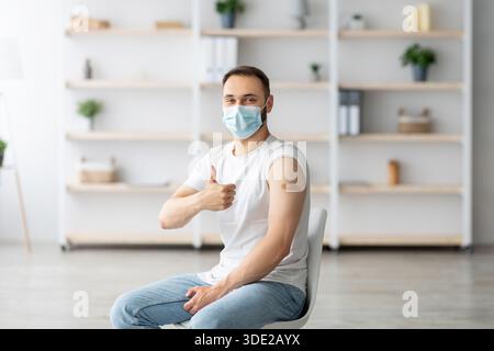 Young guy in face mask having plaster bandage on his arm after getting covid-19 vaccine injection, showing thumb up Stock Photo