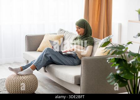 Young Arab Woman in Hijab Focused on Laptop While Relaxing at Home Stock Photo