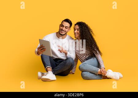 Arab Couple With Digital Tablet Relaxing On Floor Over Yellow Studio Background Stock Photo