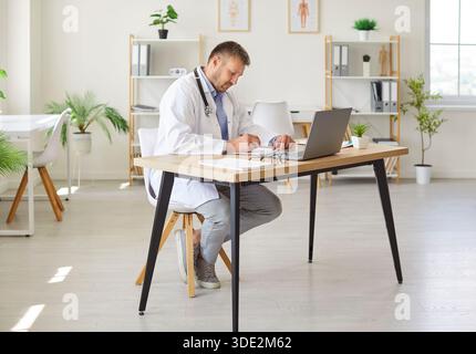 Doctor in white medical uniform working in office using laptop writing in his notepad Stock Photo