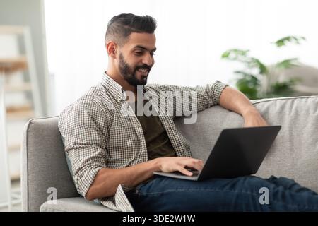 Freelancing, online job or studies. Cheerful young Arab man sitting on couch with laptop, working or learning from home Stock Photo
