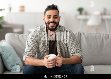Young smiling Arab guy drinking hot aromatic coffee on sofa at home, copy space Stock Photo