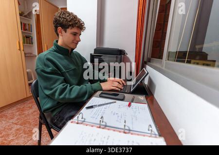 Youthful student focused on his studies, tapping away on a sleek digital tablet keyboard in a room with a view of the city through the window Stock Photo