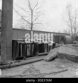 Monochrome archive photograph from the 1980s Soviet era depicts a technical utility yard, likely part of an electrical substation or maintenance facility in Brusino, Donetsk region, Ukraine USSR. The image features rows of large metal equipment housings, resembling electrical switchgear cabinets or industrial units, stored outdoors alongside pipes and concrete blocks. A brick wall encloses the site. This scene highlights the industrial infrastructure supporting the resort settlement and local logistics in the peaceful Donbass before the war, focusing on technical maintenance and engineering. Stock Photo