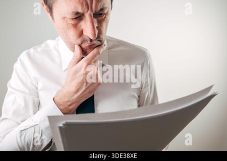 Businessman frowning deeply while holding and reading a thick stack of papers, hand thoughtfully on chin, for financial worry, bad news, contract revi Stock Photo