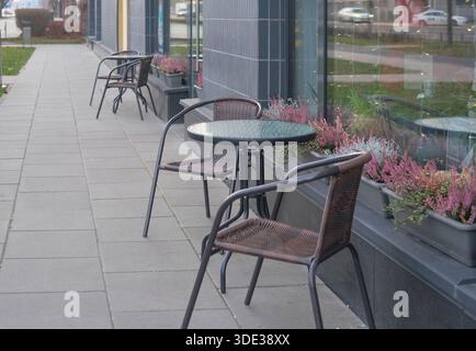 Perspective view of metal café tables with glass tops and wicker chairs on concrete paving next to a building façade, grass verge and street beyond. Stock Photo