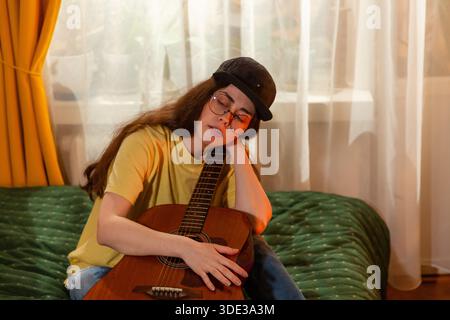Pensive Caucasian young woman student learning guitar indoors. Sitting on bed during music practice. Education and creative growth. Mid shot. Stock Photo