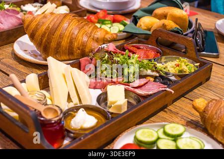Gourmet brunch tray with croissant, cheese, cold cuts, vegetables and spreads on a wooden table. Stock Photo