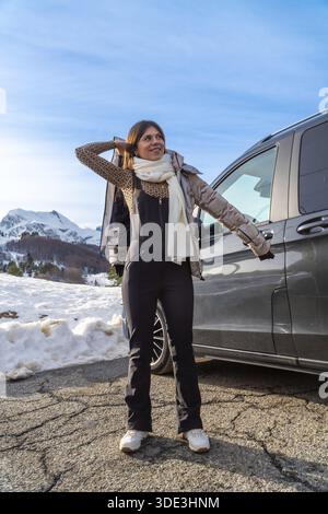 Young woman standing on a snowy mountain road next to a dark vehicle, wearing winter clothes including a leopard print top and black jumpsuit, looking Stock Photo
