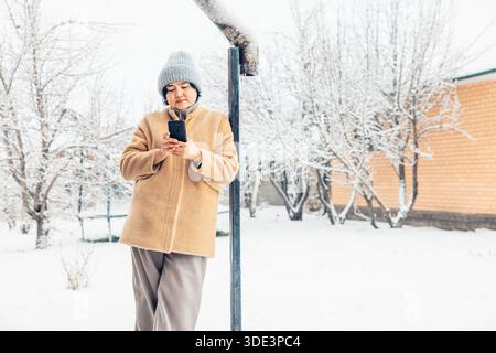 young Asian woman with smartphone in a snowy landscape. She wears a beige coat and a gray beanie. Snow-covered trees and a wooden house are in the bac Stock Photo
