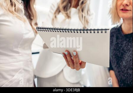A group of three women discussing skin care treatments. One woman holds a spiral notebook with before-and-after photos. The setting is clean and steri Stock Photo