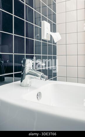 A modern bidet with a chrome faucet in a tiled bathroom. The bidet features a sleek design and is positioned against a backdrop of dark and light tile Stock Photo