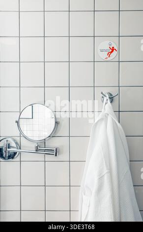 A modern bathroom with white ceramic tiles. A round mirror is mounted on the wall. A white bathrobe hangs on a hook, adding a touch of comfort. Stock Photo