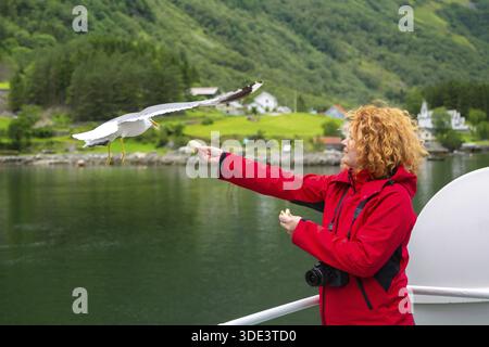 Young woman feeding seagulls flying over the ferry boat. Woman in red jacket traveling on ferry boat and feeding seagull. Woman tourist on deck of a f Stock Photo