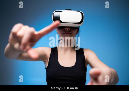 Young woman wearing VR headset with arms extended, immersive virtual reality interaction in blue studio lighting Stock Photo
