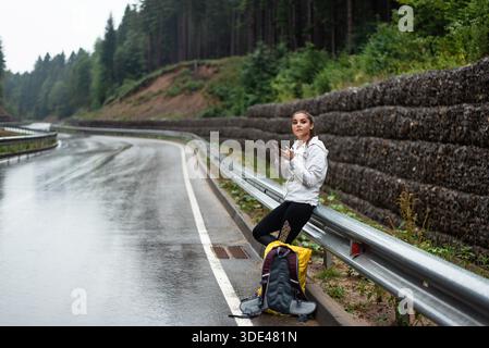 Young woman with backpack checking her phone on a wet mountain road Stock Photo