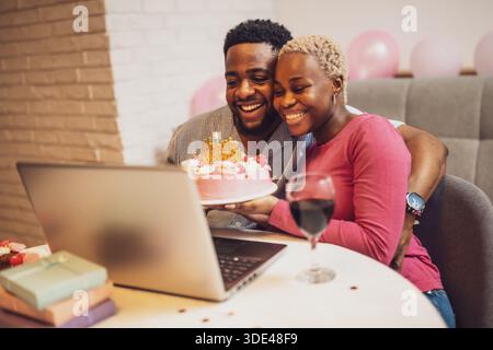 Happy young couple celebrating woman's birthday at home. They are having video call with family. Stock Photo