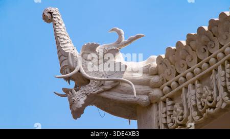 A detailed dragon head relief carved in stone adorns a traditional roof under a blue sky, showcasing mythological craftsmanship. Stock Photo