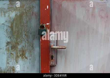 Old padlock securing a red and grey metal door, showing rust, wear, and industrial security Stock Photo