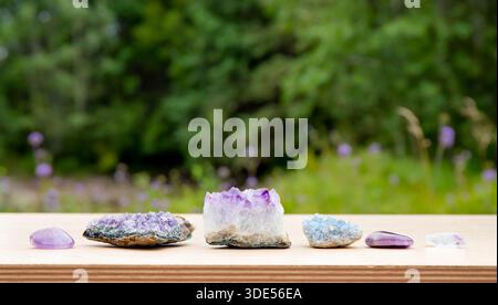 Different raw natural crystal geode clusters, amethyst, celestite, quartz on wood table with blurred wild green nature on background. Stock Photo