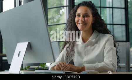 Ethnic business woman working on computer in office smiling looking at camera Hispanic businesswoman typing pc technology employee communicates with Stock Photo