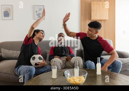 Diverse male friends sitting on sofa in matching tees, high-fiving with soccer ball, chips, bottles Stock Photo