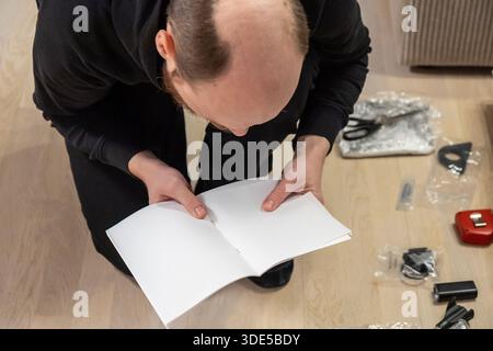 Person assembling a bathroom faucet while following an instruction manual. Components and tools, including scissors, are scattered on a wooden floor d Stock Photo