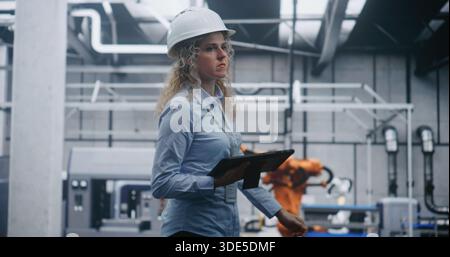 Female Industrial Specialist Wearing Hard Hat and Using Tablet to Monitor Operations Inside High Tech Factory With Robotic Arms. Concept of Automation, Efficiency, and Future of Manufacturing. Stock Photo