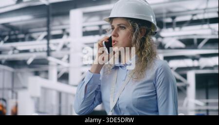 Female Engineer Wearing Hard Hat Walks Across Production Workshop and Speaks on Mobile Phone, Supervising Processes in High Tech Factory Environment. Modern Industry, Leadership and Workflow Control. Stock Photo