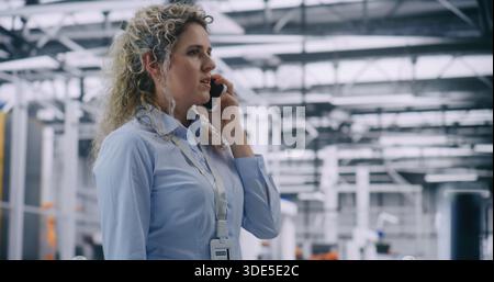 Adult Woman, Factory Supervisor, Responds Incident by Speaking Intensely on Smartphone in High Tech Manufacturing Facility. Concept of Mobile Crisis Management and Immediate On Site Decision Making. Stock Photo