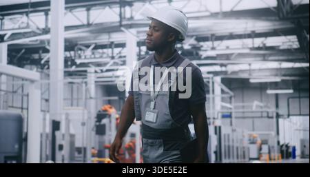 African American Male Factory Supervisor Walking Across High Tech Manufacturing Facility, Using Tablet Check Production Data, Assess Machinery Performance and Oversee Automated Industrial Operations. Stock Photo