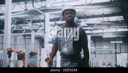 African American Male Factory Supervisor Walking Across High Tech Manufacturing Facility, Using Tablet Check Production Data, Assess Machinery Performance and Oversee Automated Industrial Operations. Stock Photo