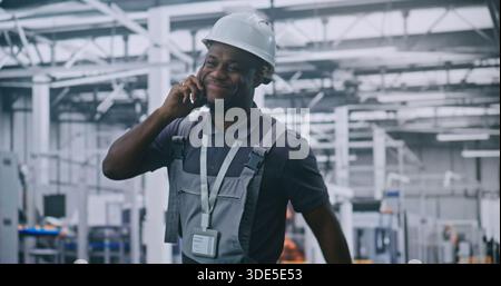 Industrial Male Engineer Walks Through High Tech Factory Floor While Talking on Smartphone and Smiling, Discussing With Colleague. Concepts Smart Manufacturing, Remote Coordination, and Industry 4.0. Stock Photo
