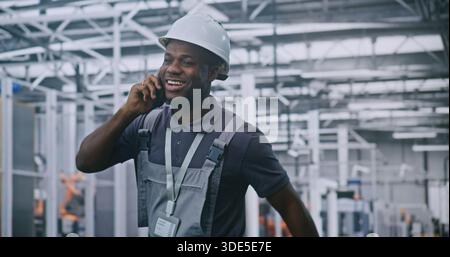 Industrial Male Engineer Walks Through High Tech Factory Floor While Talking on Smartphone and Smiling, Discussing With Colleague. Concepts Smart Manufacturing, Remote Coordination, and Industry 4.0. Stock Photo