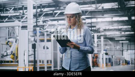 Focused Female Engineer Wearing Safety Helmet Reviews Operational Data on Digital Tablet Inside High Tech Manufacturing Facility. Concept of Digital Transformation, Modern Industrial Workforce. Stock Photo