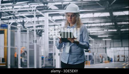 Professional Female Engineer in Hard Hat Checks Production Data on Tablet While Walking Through Modern Automated Factory. Concept of Smart Manufacturing, Industry 4.0, Digital Control and Efficiency. Stock Photo