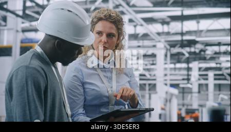 Two Specialists Solve Technical Issue in Vast Industrial Plant. Woman Points Area Concern on Factory Floor, Using Tablet For Data Reference. Problem Solving and Operational Excellence in Industry 4.0. Stock Photo