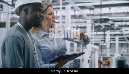 Two Specialists Solve Technical Issue in Vast Industrial Plant. Woman Points Area Concern on Factory Floor, Using Tablet For Data Reference. Problem Solving and Operational Excellence in Industry 4.0. Stock Photo