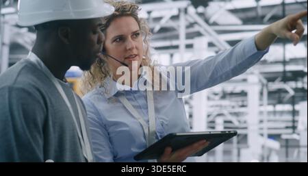 Technical Team Performing on Site Inspection in Modern Automated Factory. Female Manager Uses Tablet Review Digital Schematics While Guiding Male Engineer. Mobile Technology Integration in Industrial Audit. Stock Photo