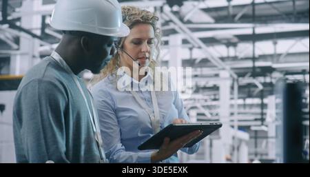 Two Specialists Solve Technical Issue in Vast Industrial Plant. Woman Points Area Concern on Factory Floor, Using Tablet For Data Reference. Problem Solving and Operational Excellence in Industry 4.0. Stock Photo
